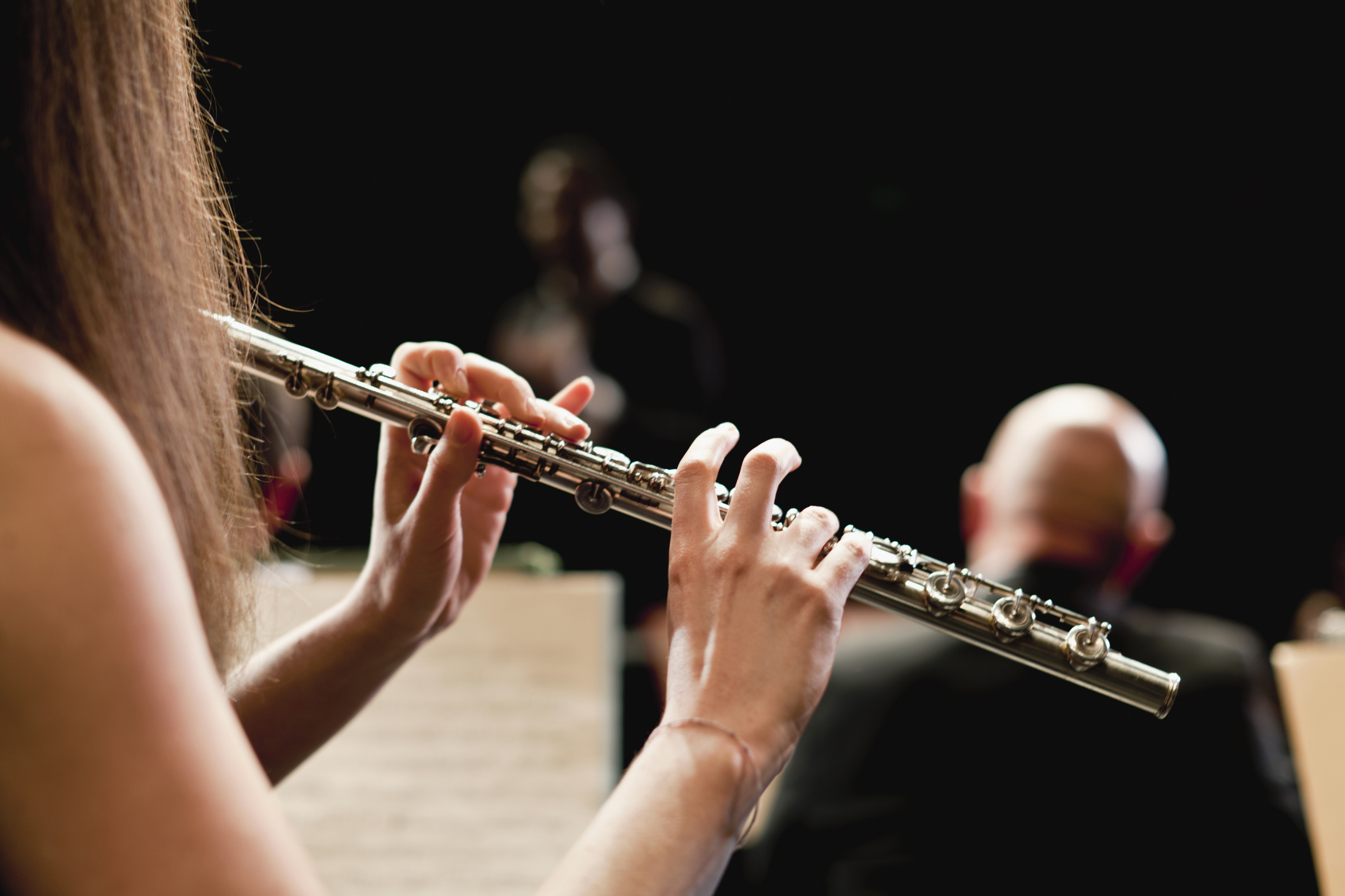 Close-up of a person playing a flute with sheet music and other musicians in the background