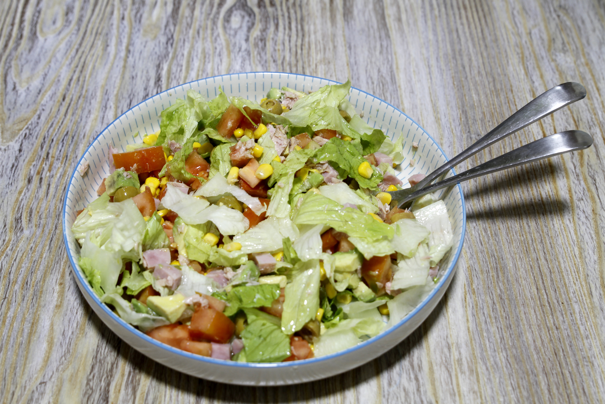 A bowl of mixed salad with lettuce, tomatoes, corn, and diced ham, with tongs on a wooden surface