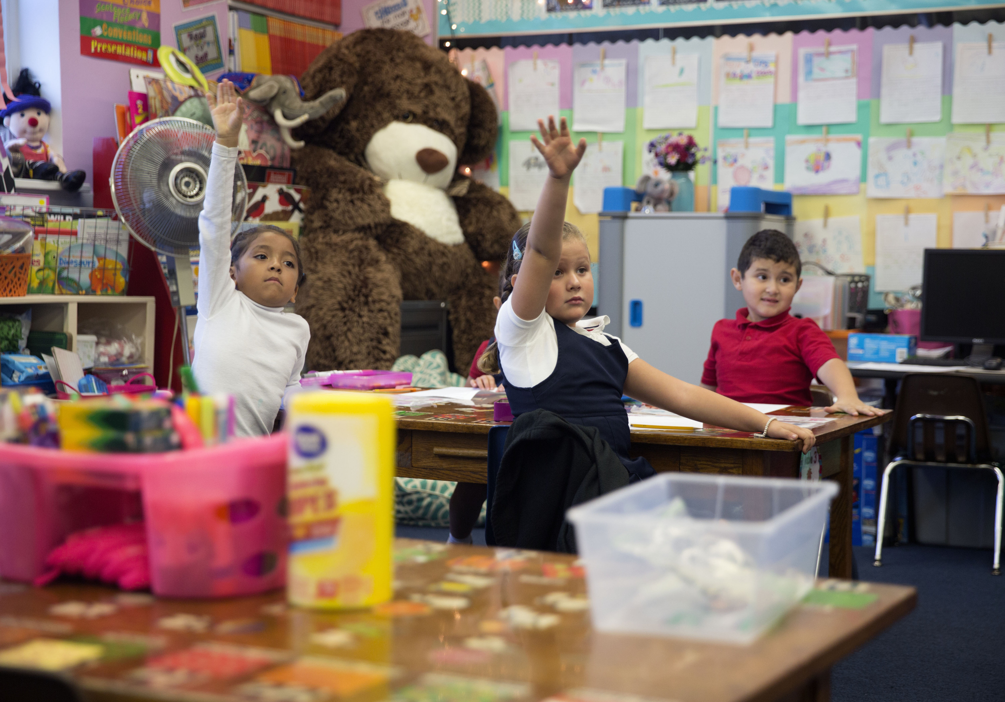 Three children in a classroom setting with their hands raised, looking eager to answer a question