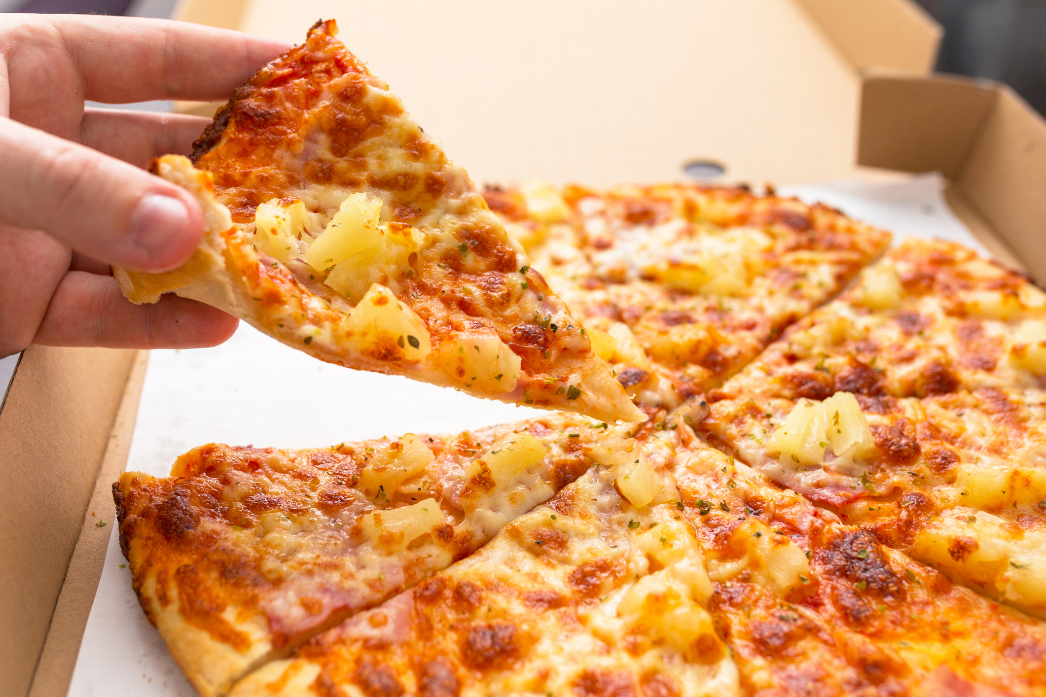 Hand holding a slice of pizza with pineapple and ham toppings, taken from an open pizza box