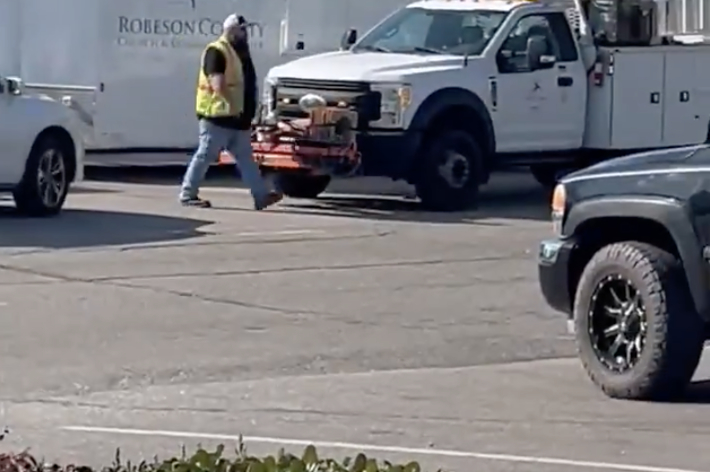 Person in high visibility vest walking across a parking lot, with trucks and trailers in the background