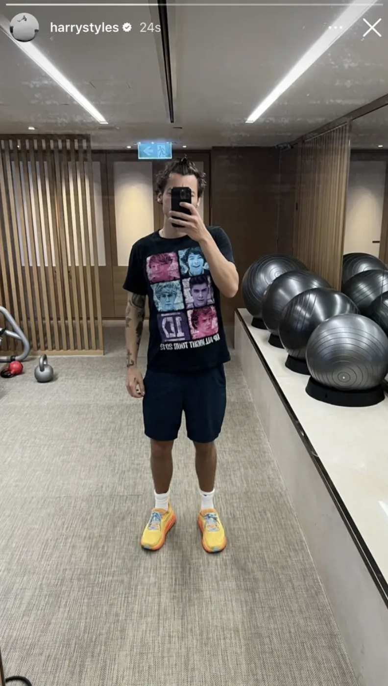Harry in 1D tee and shorts taking a mirror selfie in a gym, wearing sneakers