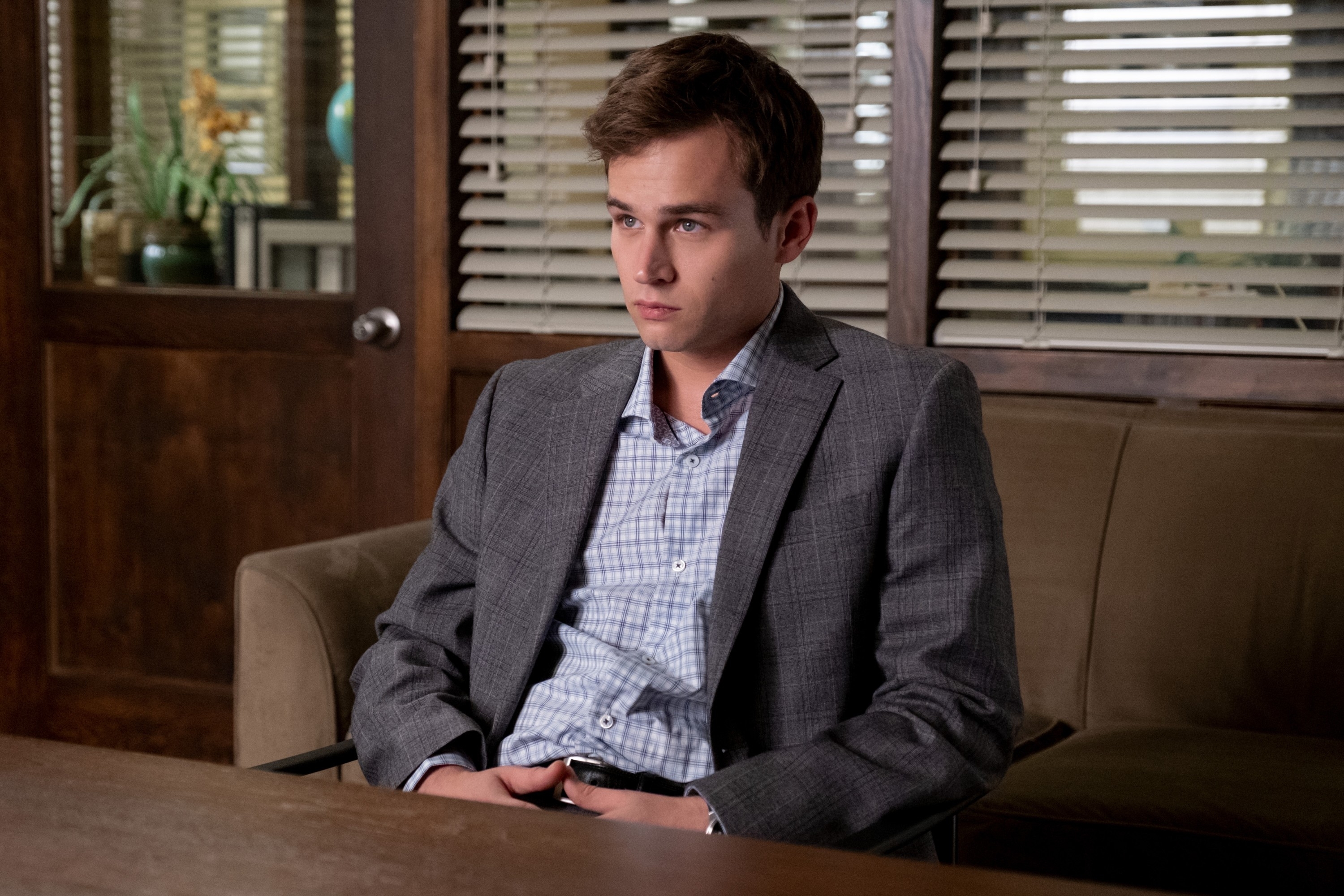 Character from a TV show sitting in an office wearing a suit and checkered shirt