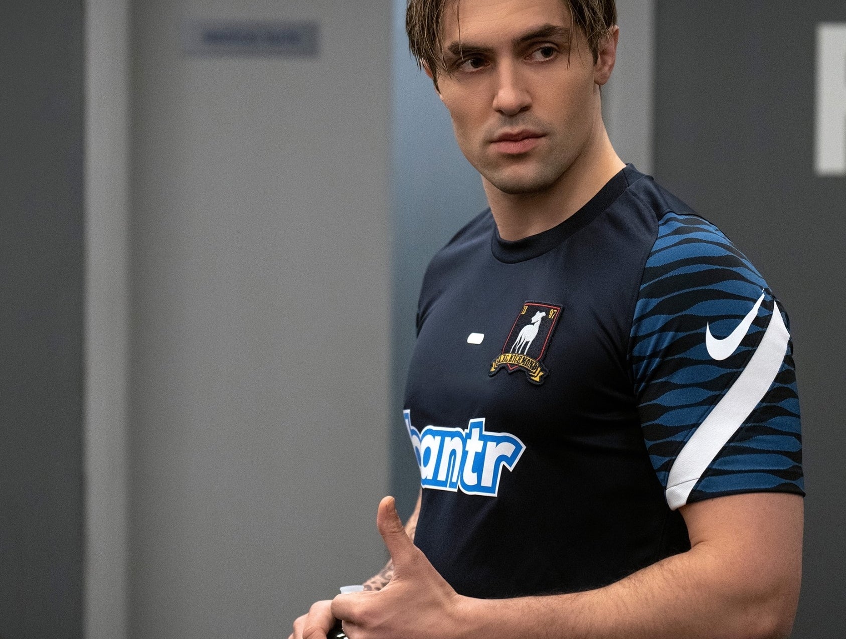 TV character in a soccer jersey stands in an office setting, looking off-camera