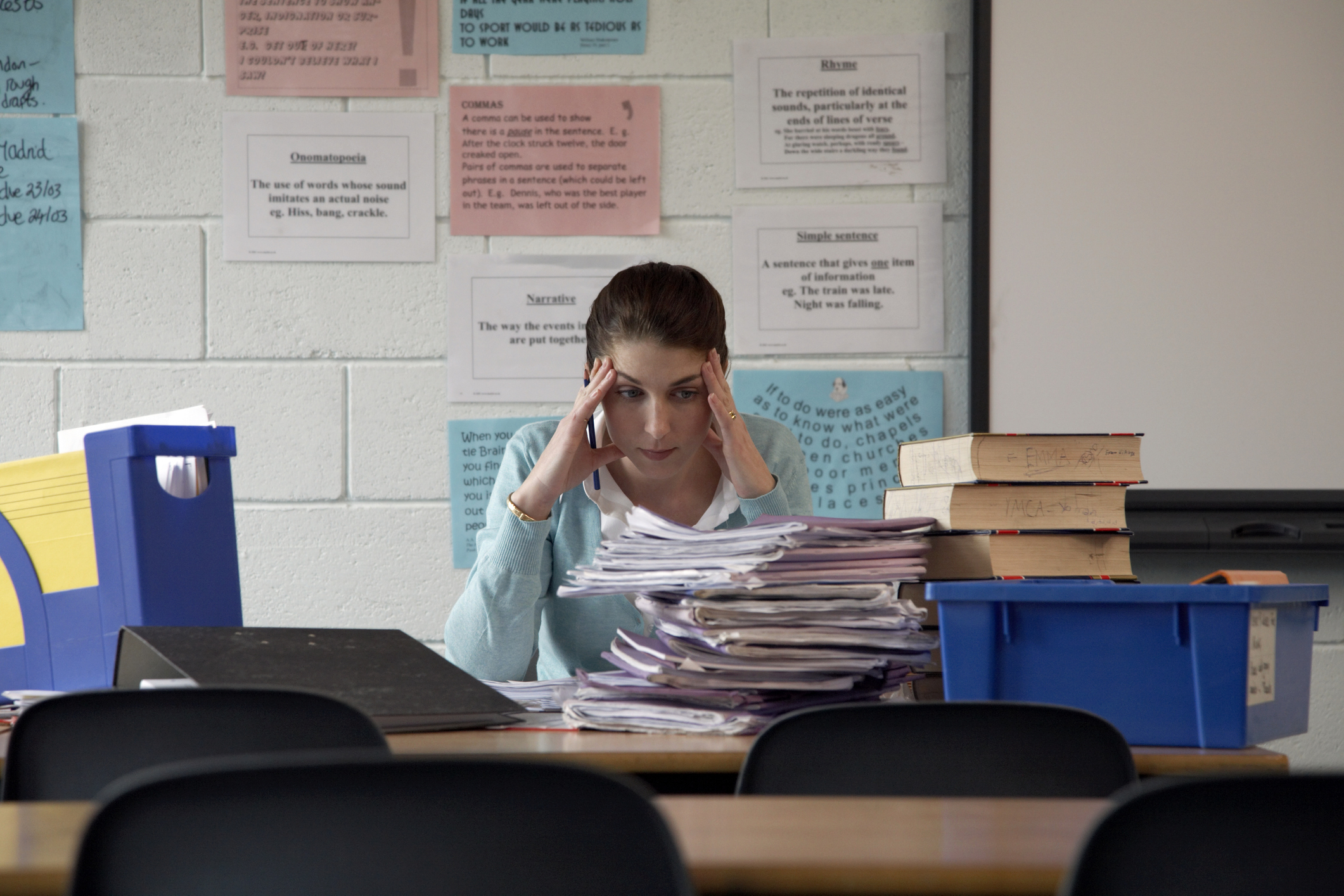 Teacher sitting at a desk with a stack of papers, appearing stressed or thoughtful