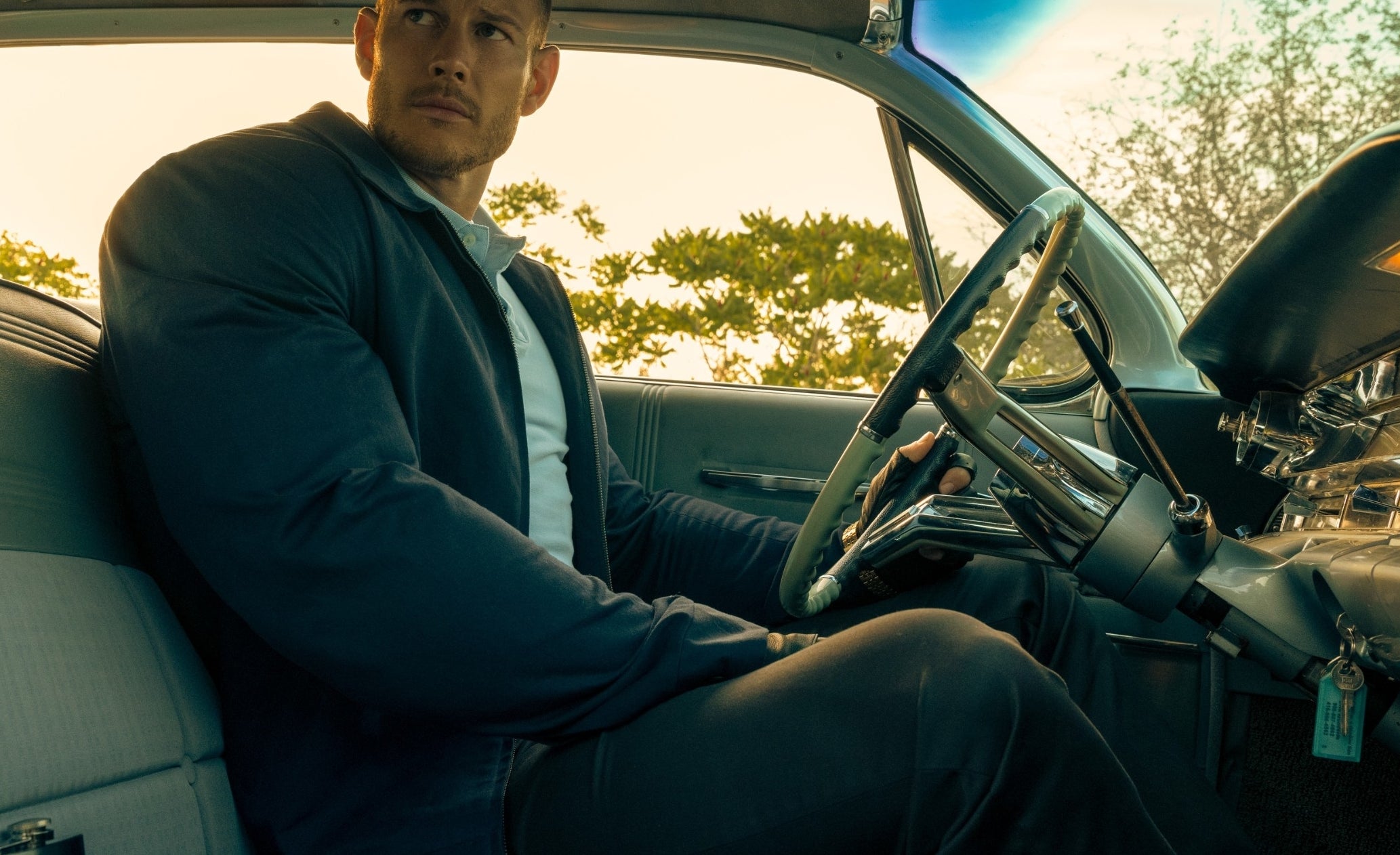 Man in a suit sitting inside a classic car with a stern expression, handgun on the seat beside him