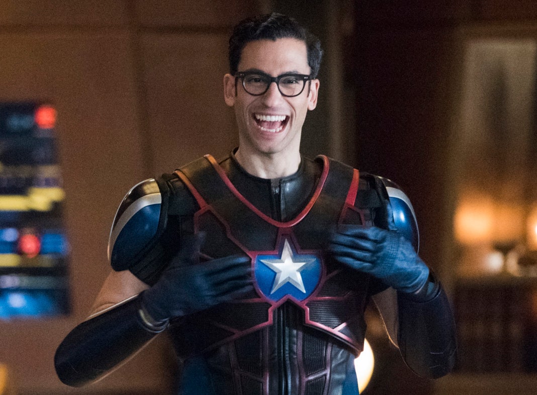 Character in superhero costume stands smiling with hands on hips in a room with large screen in the background