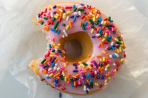 A frosted donut topped with sprinkles on wax paper with a bite taken out of it