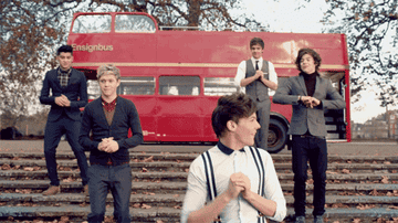 Cinco miembros de One Direction posando frente a un autobús rojo de dos pisos; algunos están de pie y otros sentados en escaleras