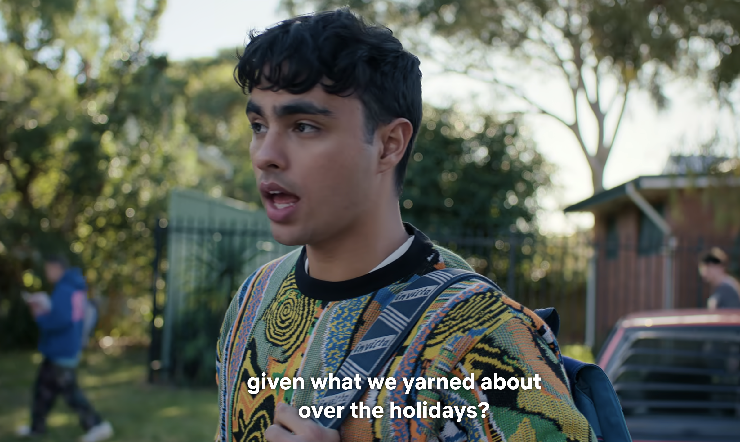 Young man with a concerned expression wearing a patterned sweater outside. Subtitles on screen