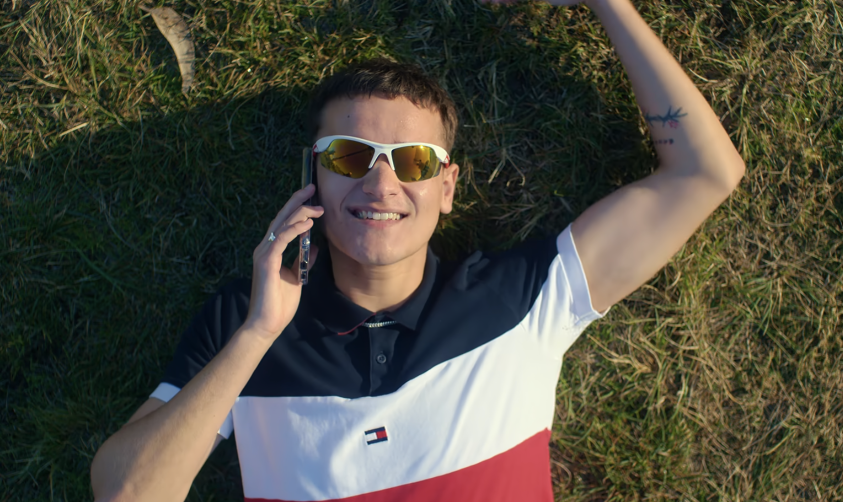 Man in Tommy Hilfiger shirt and sunglasses lying on grass, talking on phone, smiling