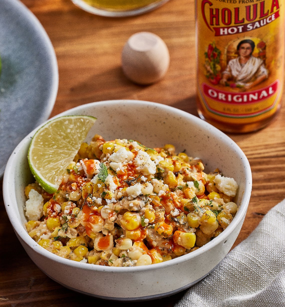 A bowl of elote with lime wedges next to a bottle of Cholula hot sauce