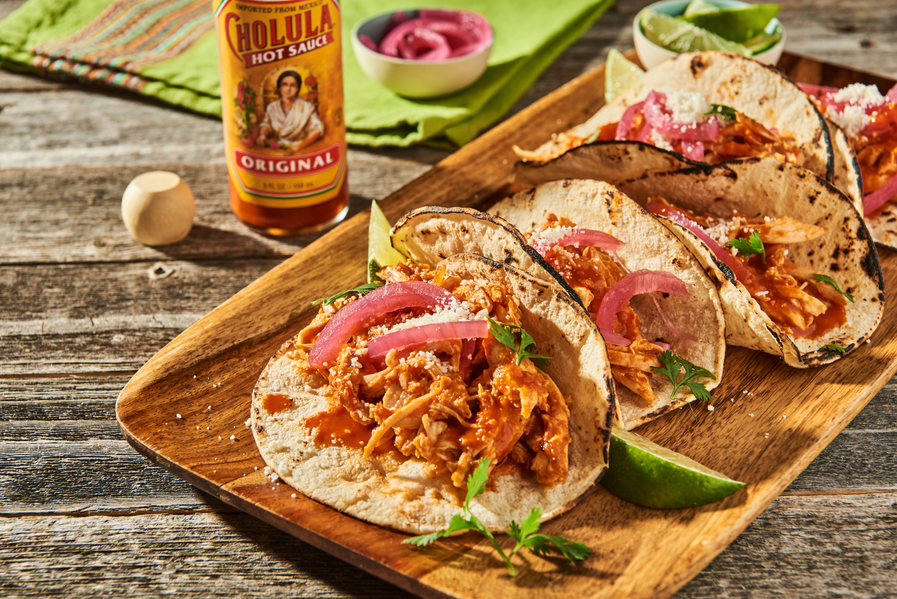 Three tacos with chicken and sliced onions on a wooden board next to a bottle of Cholula hot sauce