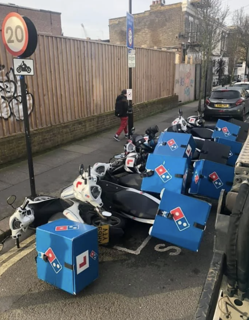 A person walks past a pile of fallen Domino’s Pizza delivery scooters on a street