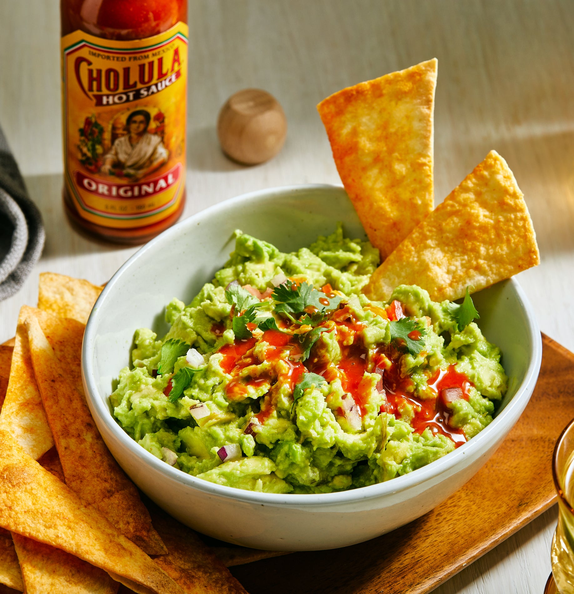 Bowl of guacamole with tortilla chips and a bottle of Cholula hot sauce on a table
