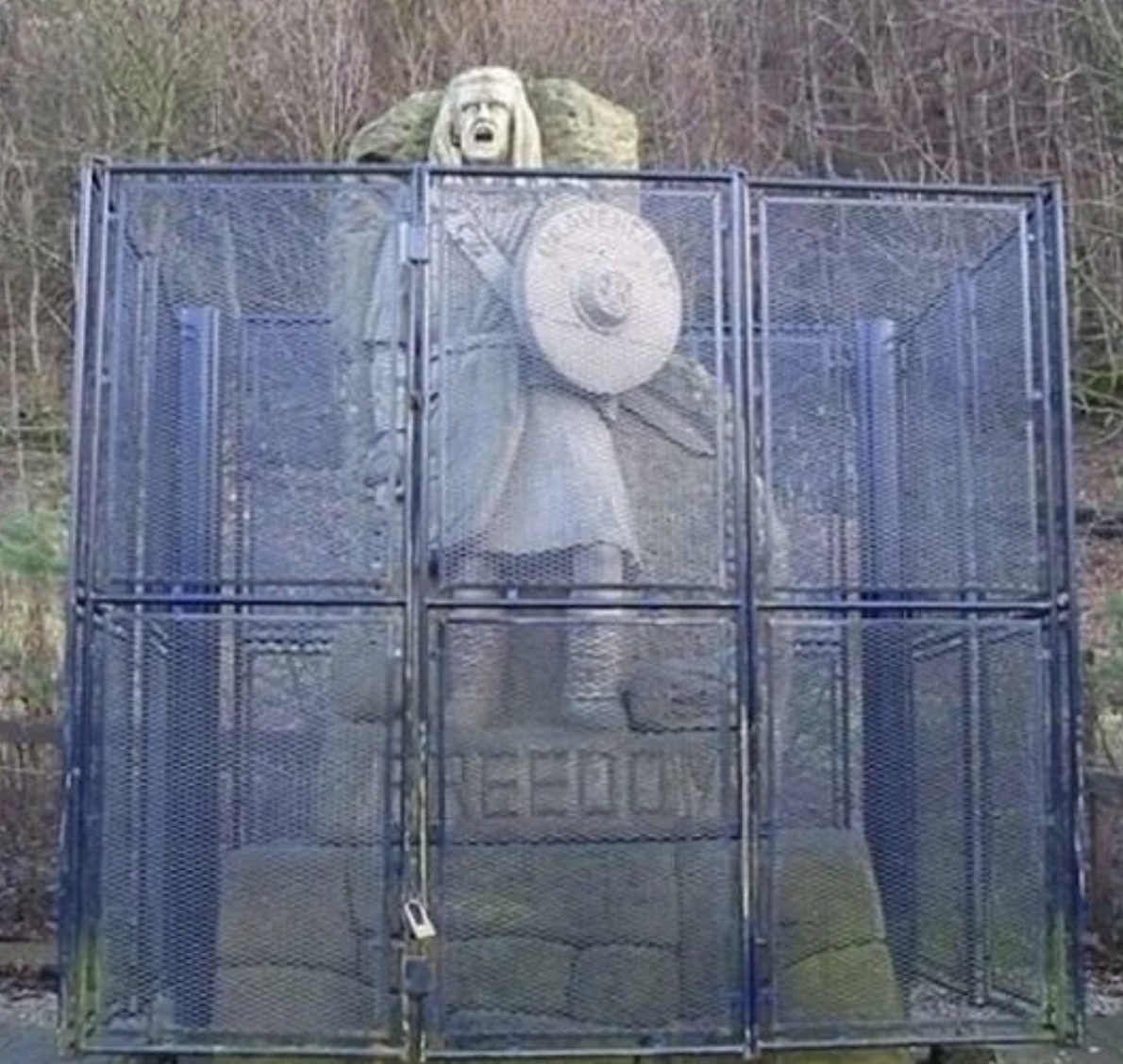 Statue of a figure with a shield and sword trapped behind a metal cage, with the word &quot;FREEDOM&quot; at the base