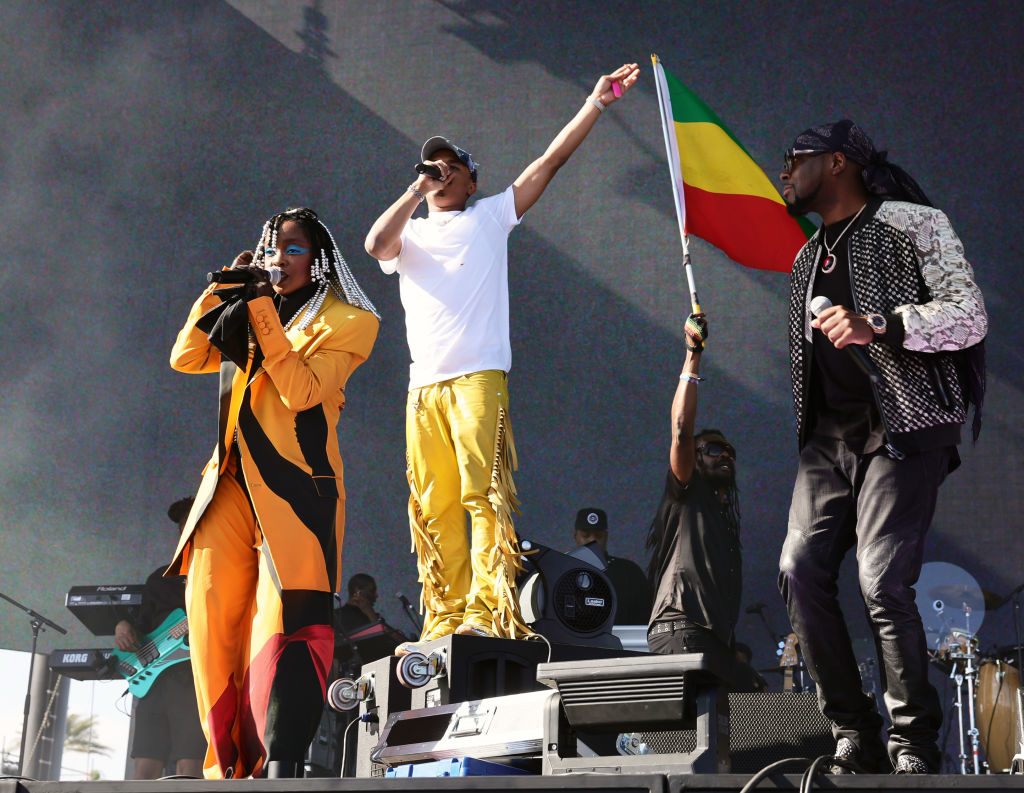 Three performers on stage, one holding a microphone, another raising a flag, with a crowd backdrop