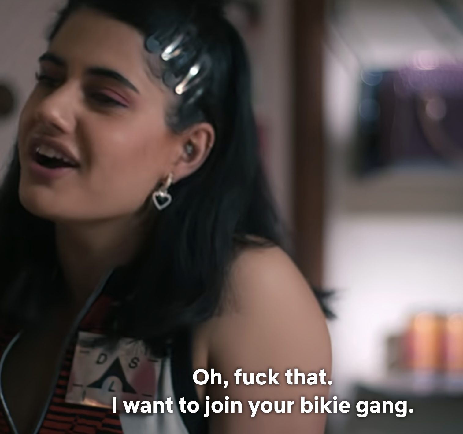 A woman with hair clips smiles and speaks, wearing a striped tank top; subtitle expresses her desire to join a &quot;bikie gang.&quot;