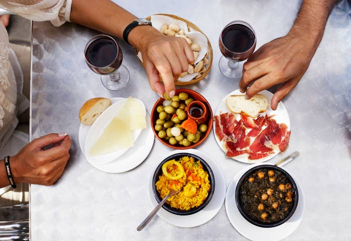 Two people dining with tapas dishes, bread, cheese, and red wine on the table. Hands visible, no faces shown