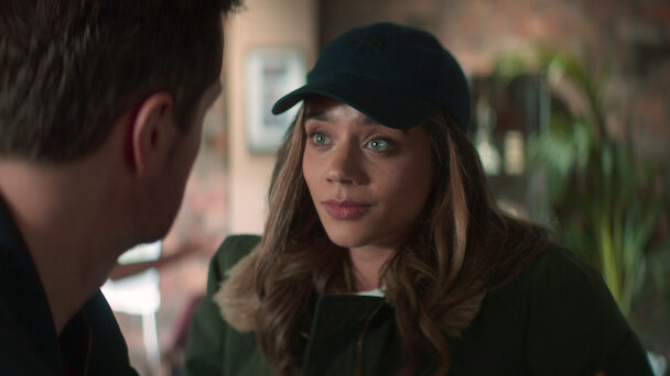 Woman in a cap facing a man, both appear to be in a serious conversation, possibly from a TV show or movie scene