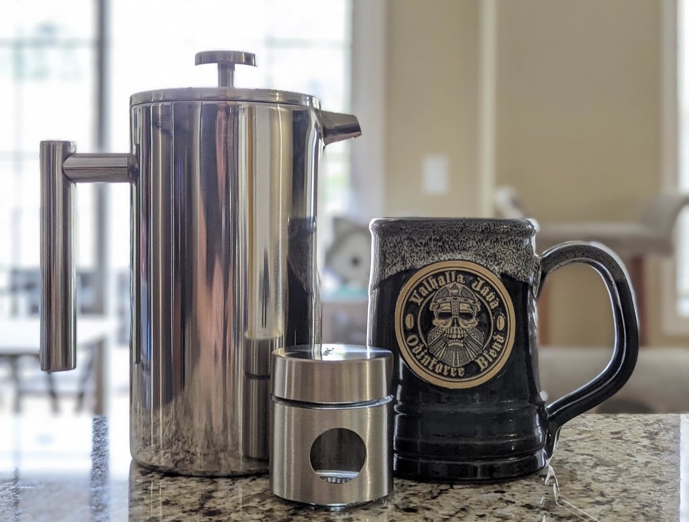 Stainless steel French press with matching container next to a ceramic mug with intricate design