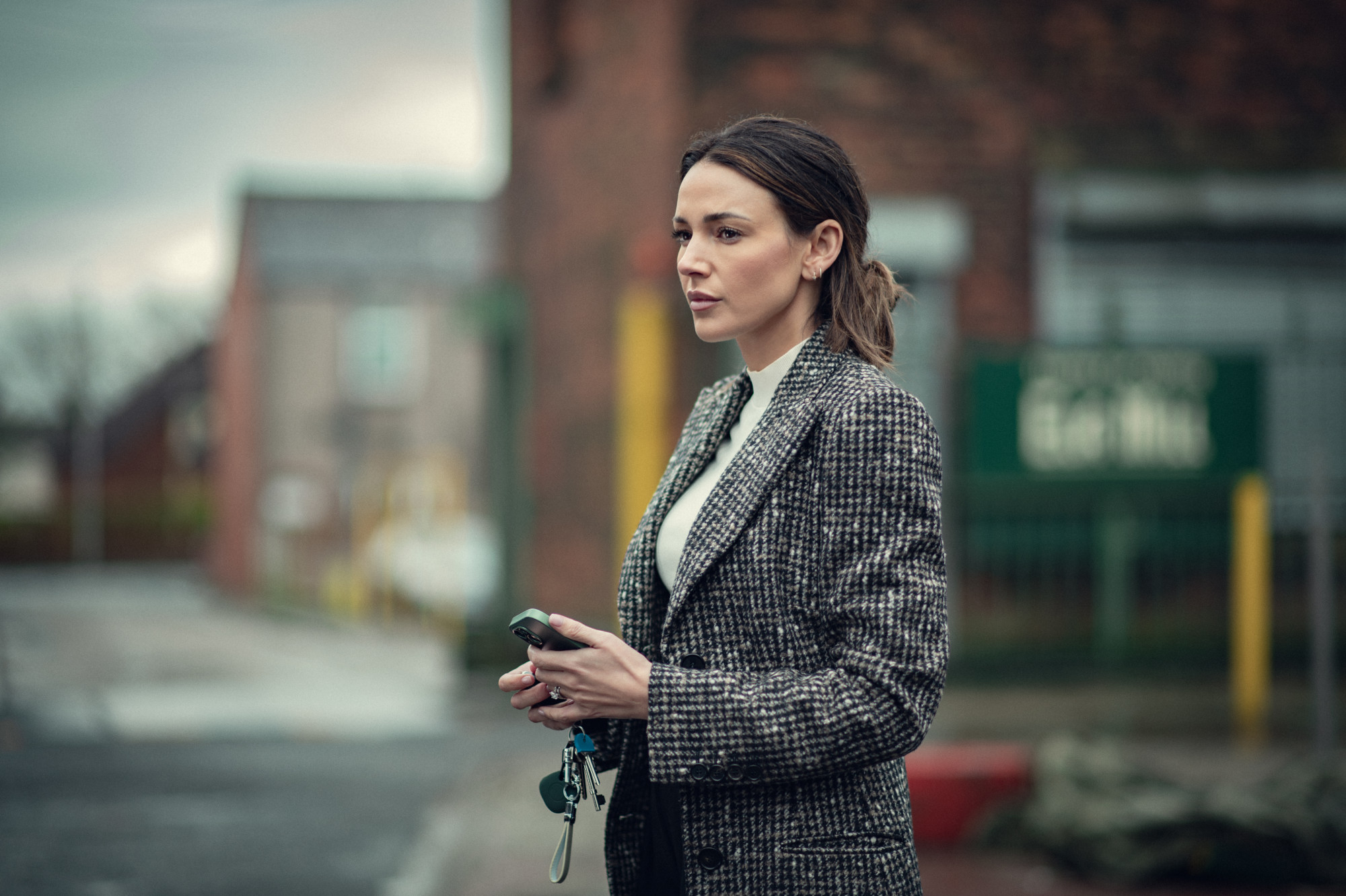 Woman in a tweed coat stands outdoors holding a phone with a focused expression