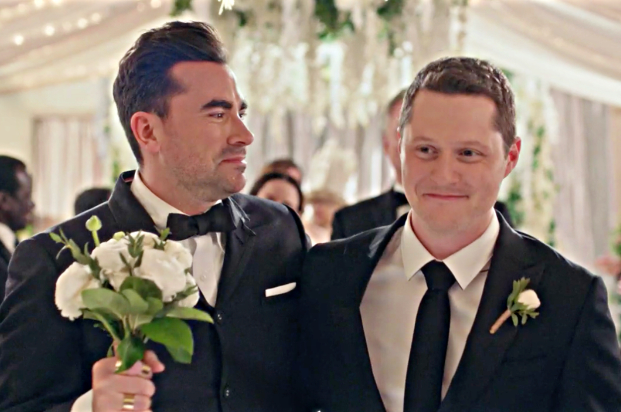 Two men in tuxedos at a wedding, one holding a bouquet, looking happy