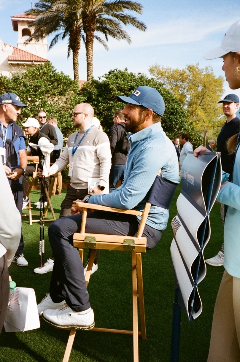 A golfer in sportswear sits on a chair outdoors surrounded by people, at a golf event
