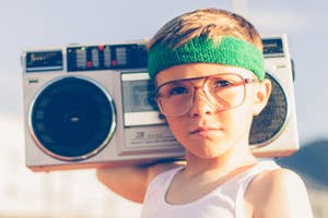 Child in headband and sunglasses holds a boombox on their shoulder, exuding a retro vibe