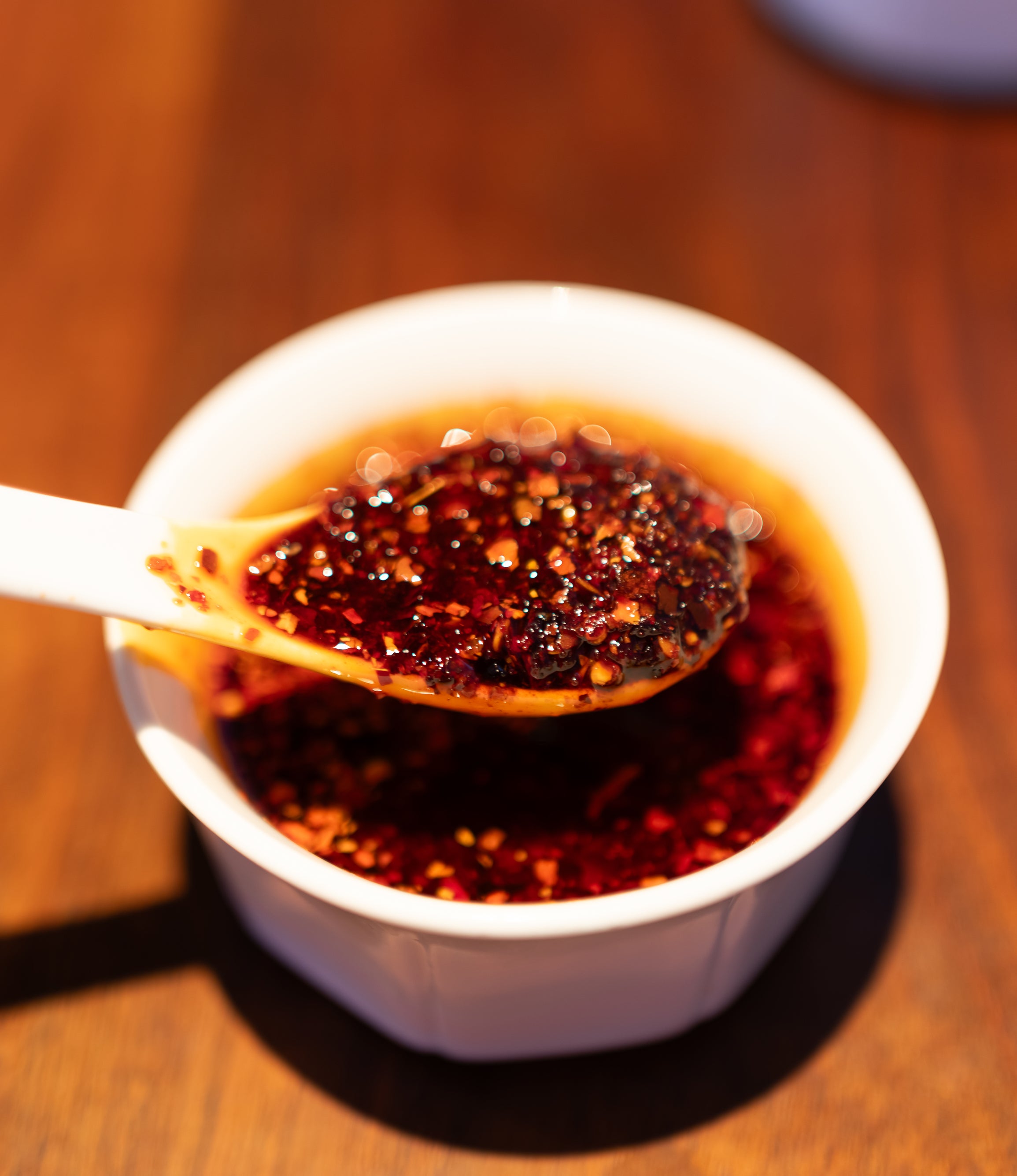 A spoon lifting chili oil with pepper flakes from a small bowl