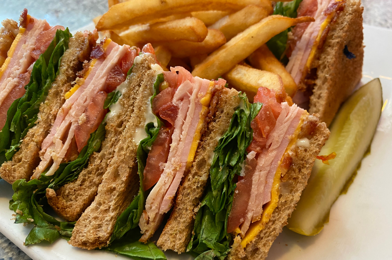 A close-up of a club sandwich with bacon, lettuce, tomato, and cheese, served with french fries and a pickle on the side