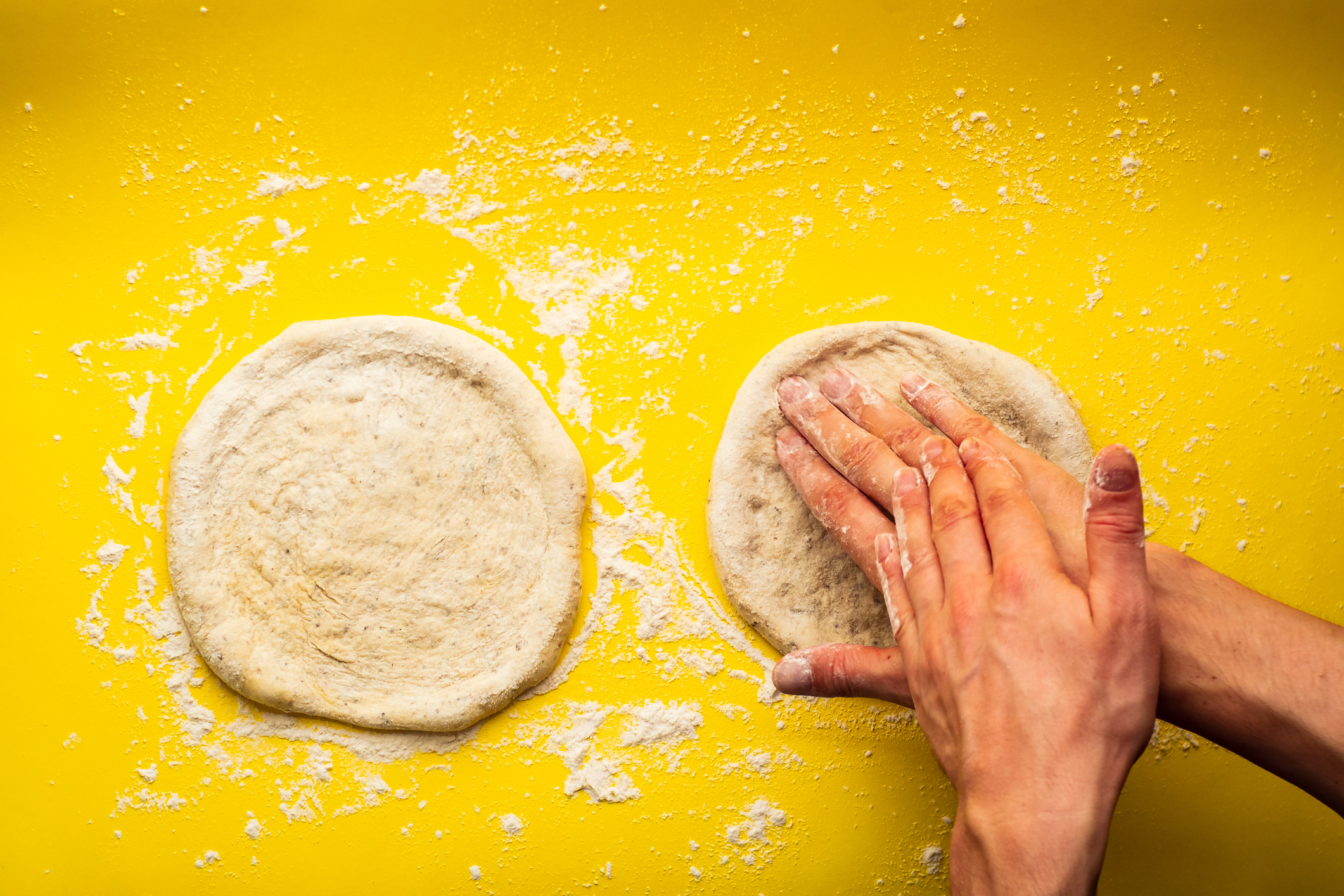 Hands pressing dough on a yellow surface, scattered with flour, for bread or pizza making