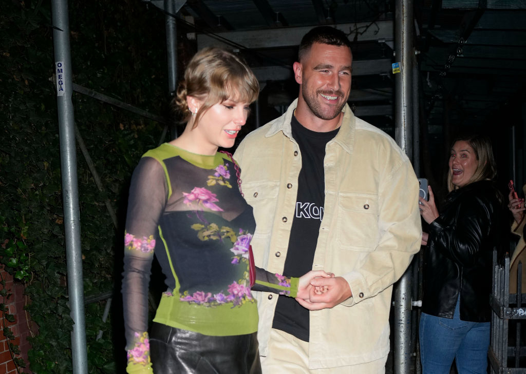 Taylor Swift in a floral dress holding hands with Travis in a jacket, both smiling