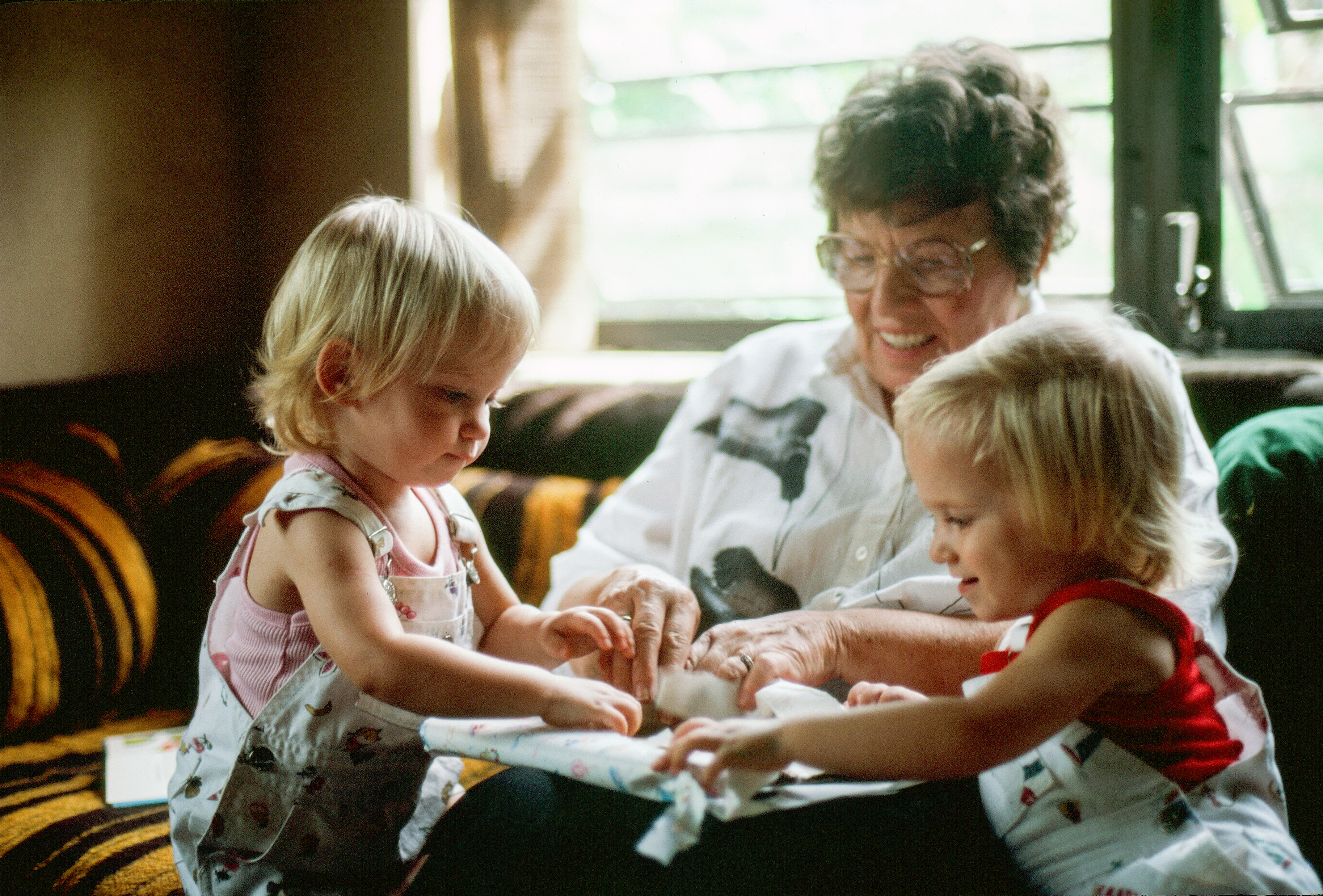 Grandmother with glasses sits with two young children who are playing with a toy