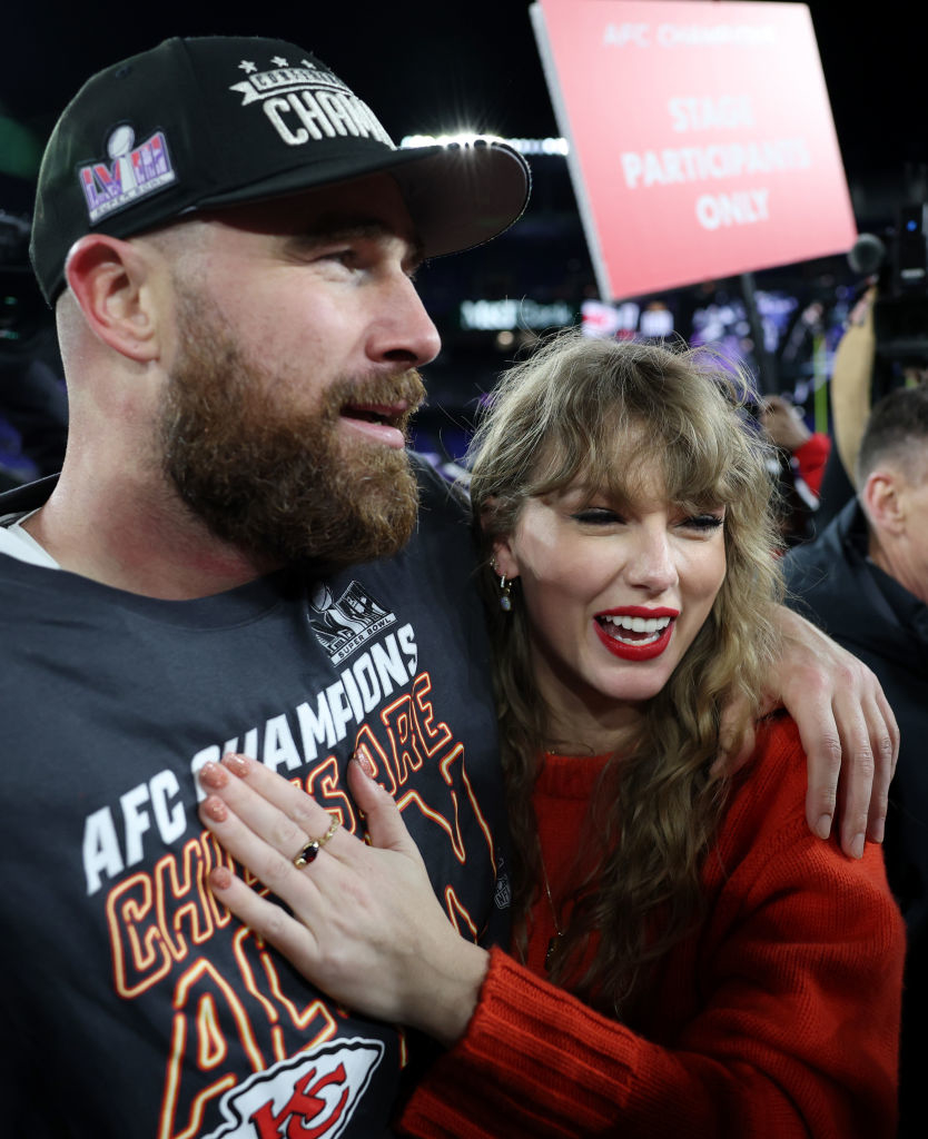 Travis Kelce in a championship shirt hugging a smiling Taylor Swift in a sweater at a sports event