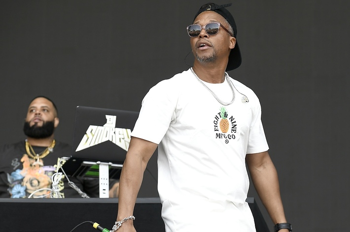 Lupe Fiasco performs on stage wearing a white t-shirt and sunglasses with a DJ in the background