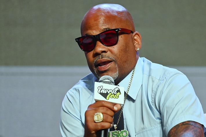 Damon Dash speaking into a microphone at an event, wearing a light blue shirt, sunglasses, and gold accessories