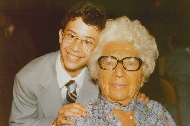Young person in formal attire stands smiling with elderly woman in a floral blouse