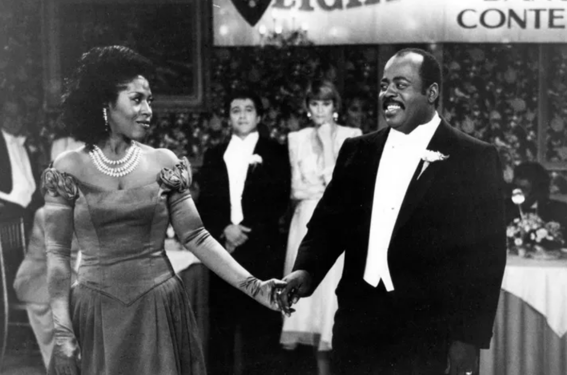 Two actors on set, woman in a vintage dress and man in a tuxedo, holding hands and smiling