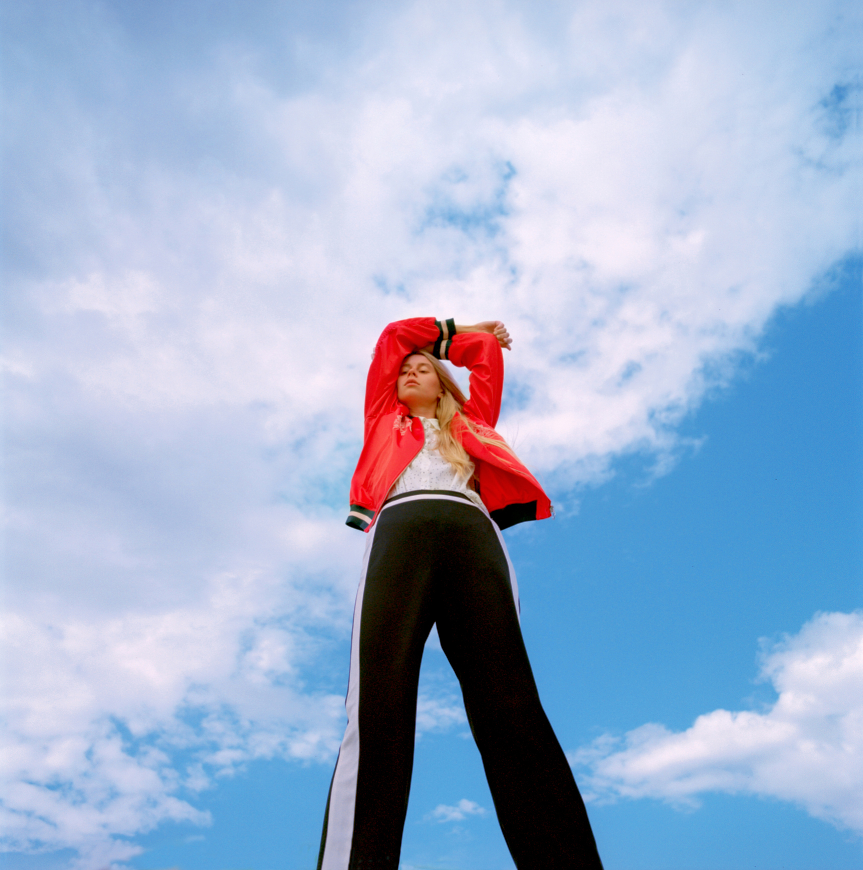 Person in a red jacket and black pants poses against a cloudy sky
