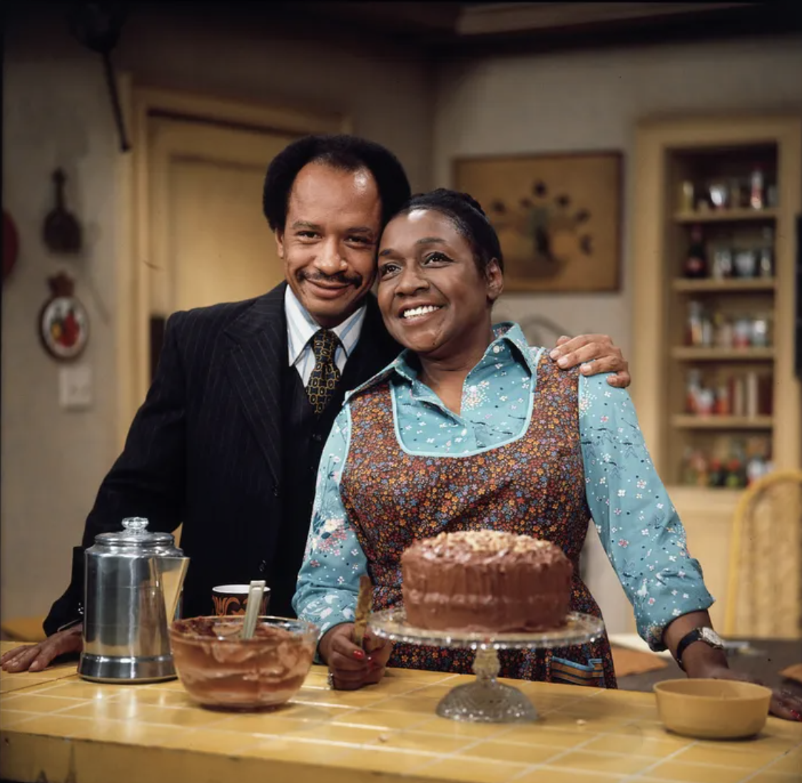 Two characters from a classic TV show in a kitchen, man in suit and woman in apron, smiling with a cake on the table