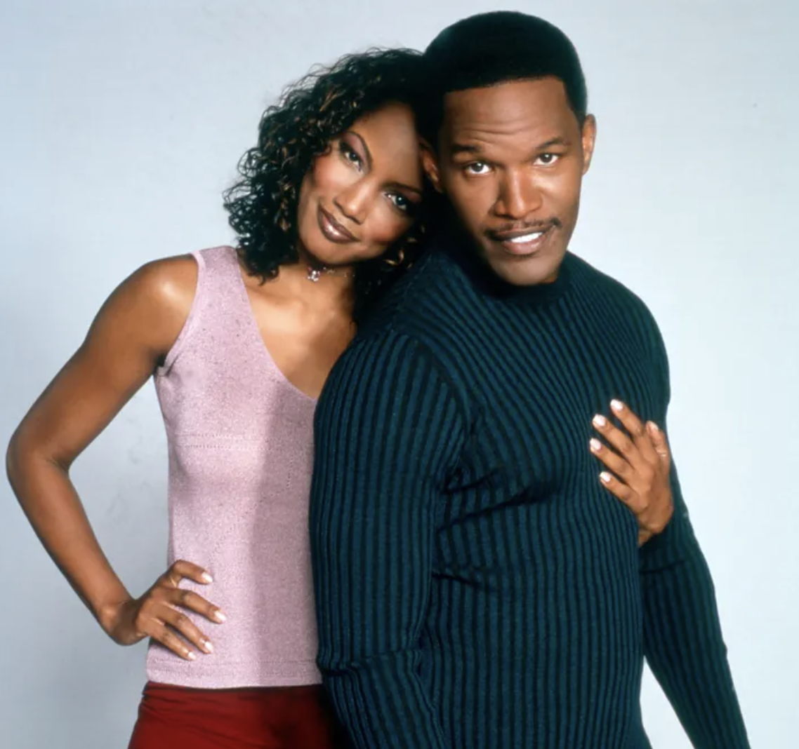 Two individuals posing together, one in a sparkly sleeveless top and red pants, and the other in a ribbed sweater