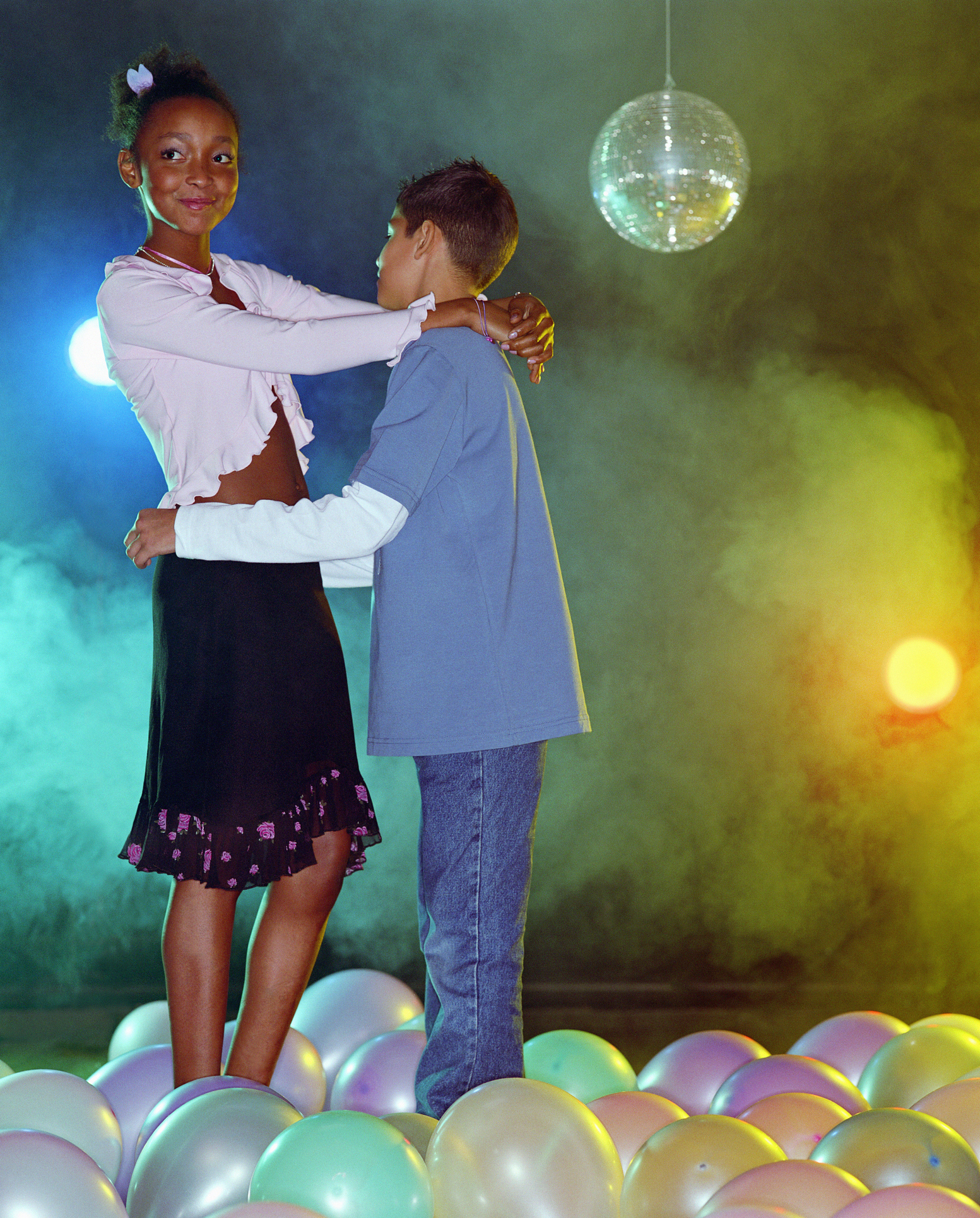 Two children dancing together at a party with a disco ball overhead and balloons on the floor