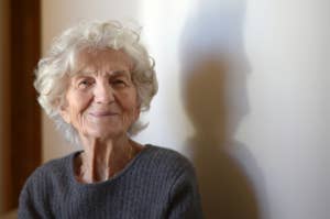 Elderly woman with a gentle smile wearing a casual sweater, with soft indoor lighting and shadow play on the wall