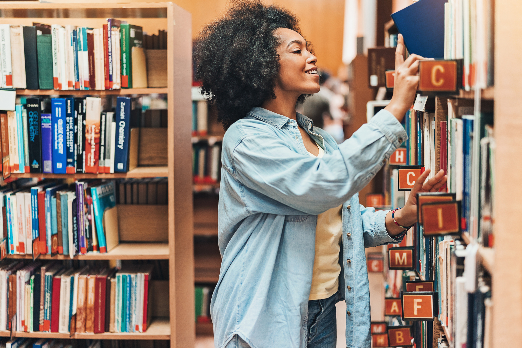 A woman is selecting a book from a library shelf, smiling. She&#x27;s in casual attire