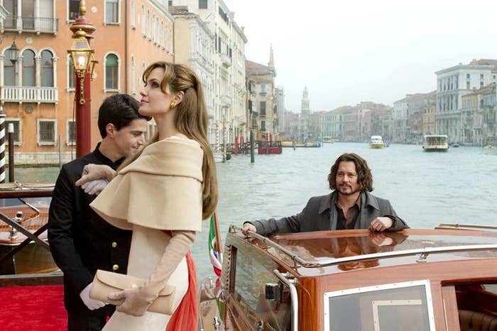 Angelina Jolie in a chic beige cape, planting a kiss on a man&#x27;s cheek, while another man stands by a boat in Venice