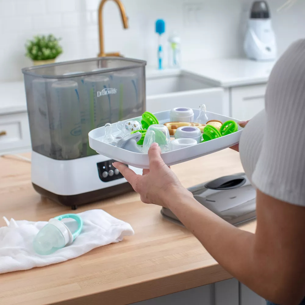Person using a bottle sterilizer with various baby items on kitchen counter