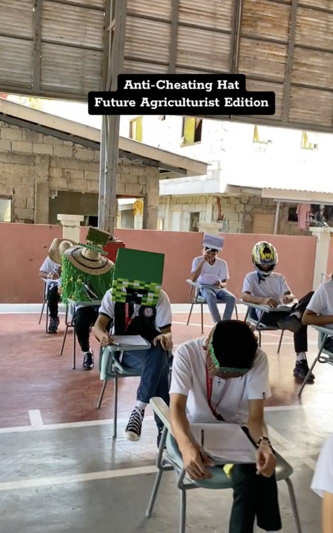 Students wearing homemade hats with extended flaps to prevent cheating during an exam, one hat styled like a grass block from Minecraft