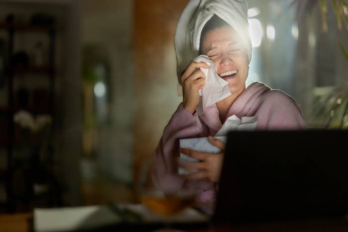 Woman in robe and towel on head laughing while wiping tear from eye, in front of laptop