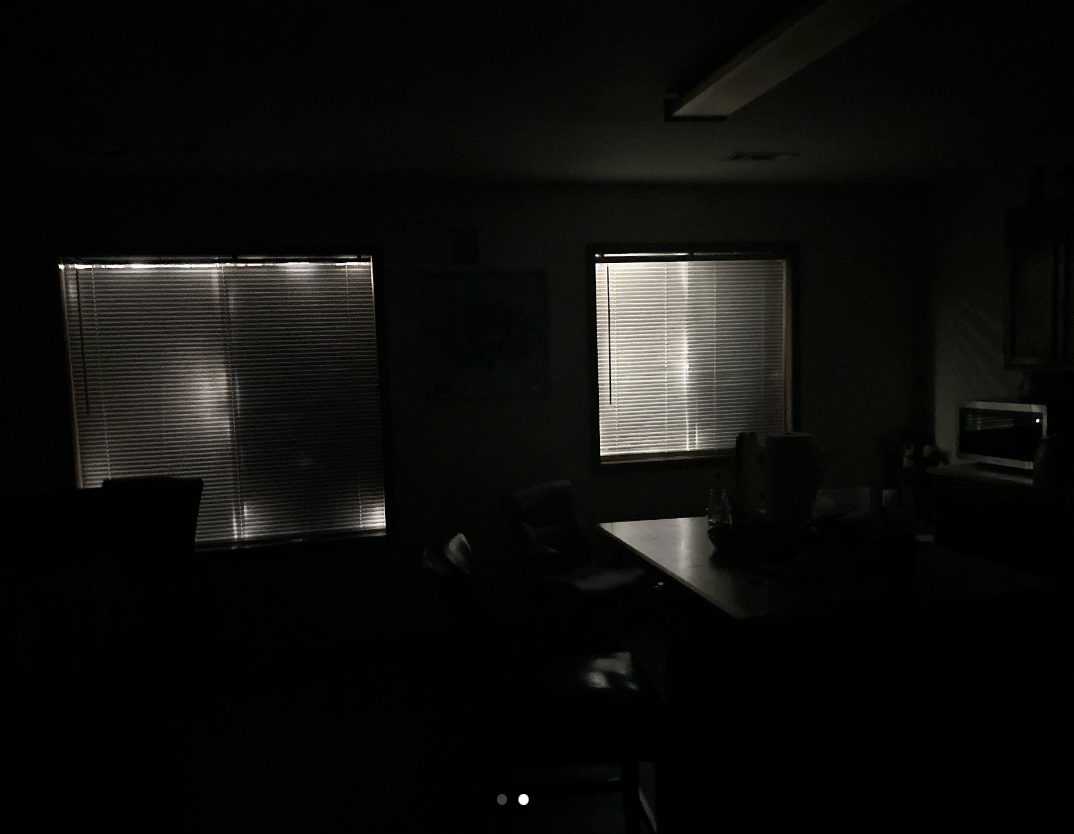 Dark room with closed blinds on windows and silhouette of a table and chair, conveying a quiet and dim setting